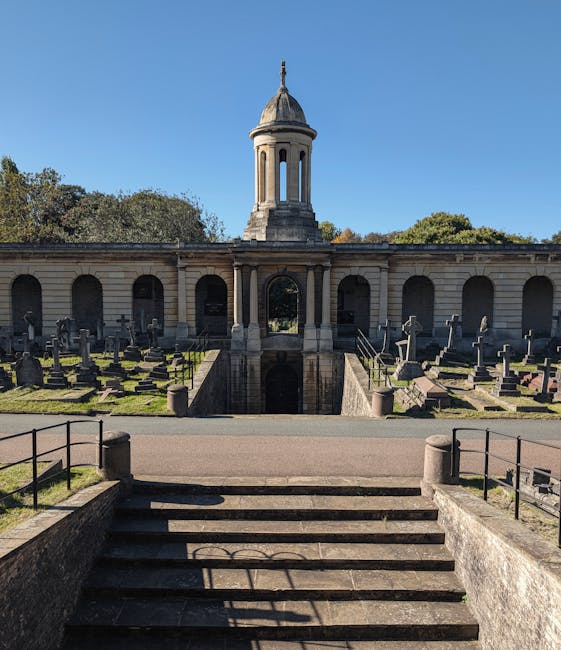 The image shows a historic cemetery with a symmetrical layout, featuring a central stone archway leading down to a burial area with rows of headstones and grave markers. Behind the arches, there is a 