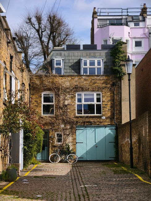 A narrow cobbled alleyway in Brompton, showing the rear of a traditional two-storey brick house with two white-framed windows on the upper floor and a smaller window on the ground floor, alongside a pair of light blue wooden garage doors. An upright bicycle rests against the brick wall near the garage, and dense ivy and foliage grow partially over the upper windows and along the side of the building. A black street lamp is mounted on a pole on the right side of the alley, and the scene is illuminated by natural daylight. This setting depicts a typical residential area where house removals, packing, and furniture transport might occur, with [COMPANY_NAME] providing professional moving services to facilitate home relocation in Brompton.