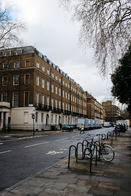 A street scene outside a multi-storey residential building with a brown brick facade, white window frames, and balconies on each level. Several large cardboard boxes and wrapped furniture are loaded onto a light-grey removal van parked on the pavement in front of the building, with the rear doors open and the loading process in progress. A person in a yellow jacket is walking nearby, assisting with the move. In the foreground, there are metal bike racks, one bicycle locked to one of them, and a sidewalk with a few pedestrians passing by. The environment is urban, with overcast sky and leafless trees on the right, indicating a late autumn or winter period. This scene shows the logistics involved in house removals, small-scale furniture transport, and packing during a home relocation, demonstrating equipment and activity typical of professional moving services such as those offered by Removal Van Brompton.
